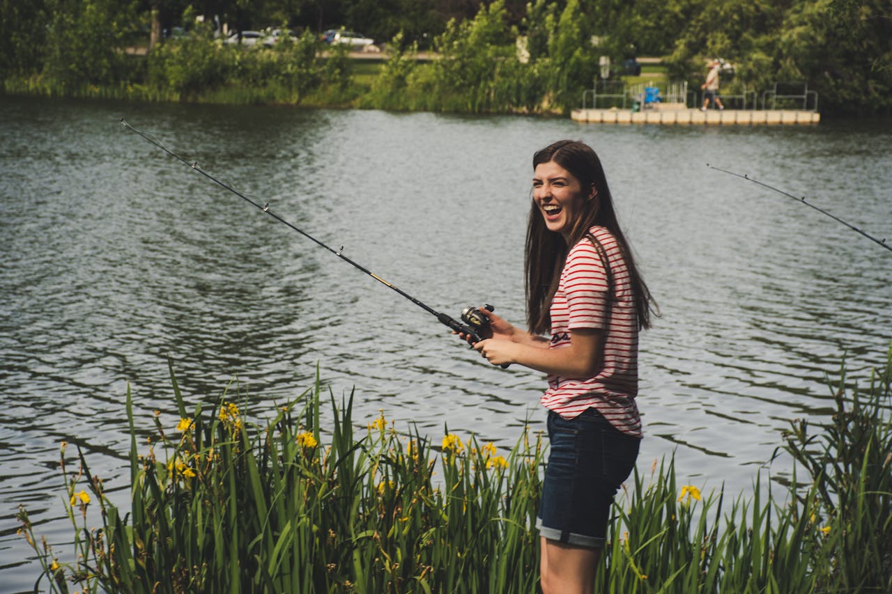 A cheerful young woman fishing by a serene lake surrounded by greenery. Perfect day outdoors.